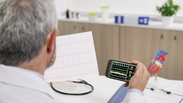 Man doctor in office analyzes remotely patient condition real time remote assistance,male medic sitting at his desk examines heart data ecg live streaming from a sick person at home 