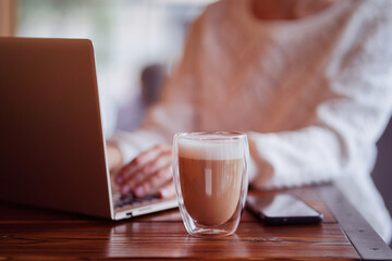 Coffee break. Cup of cappuccino on working table with a woman typing on a laptop.