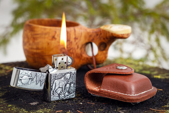 Zippo Timberwolves lighter burning with a leather case and a wooden kuksa with delicious coffee on an old stump in the spring forest. The background is blurred.