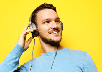 Handsome man adjusting his headphones and smiling while standing against yellow background