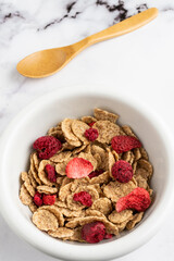 Aerial view of bowl with cereals, red berries and wooden spoon, on white marble table, in vertical, with copy space