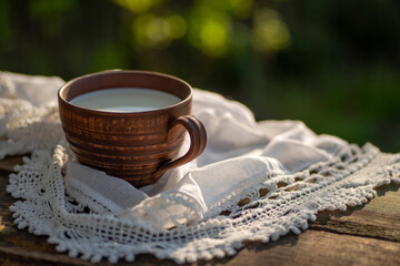 Obraz na płótnie Canvas Pottery cup with milk (kefir, yogurt, sour cream, kumis), lace tablecloth, wooden table. Outdoor picnic, breakfast, brunch, refreshments. Soft focus