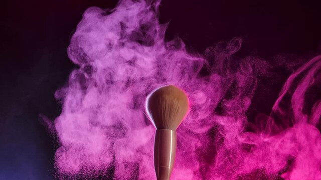 Two makeup brushes collide and cause a swirl of powder particles against a black background in pink neon light. Fine dust of the crushed powder flies beautifully in the air. Close up. Slow motion.