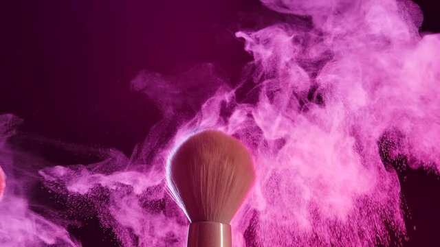 Two makeup brushes touching each other form a beautiful swirl of cosmetic powder particles. Close up of makeup brushes in dark studio in pink neon light. Slow motion.