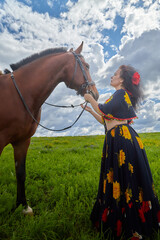 Obraz premium Beautiful gypsy girl with a horse in field with green glass in summer day and blue sky and white clouds background. Model in ethnic dress posing with farm animal