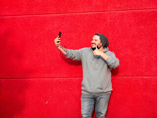 Hombre haciéndose un selfie en una pared roja vistiendo mascarilla sanitaria cumpliendo las...