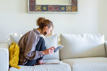 Young woman making online shopping (choosing gifts) while sitting on the sofa in a cozy home interior.