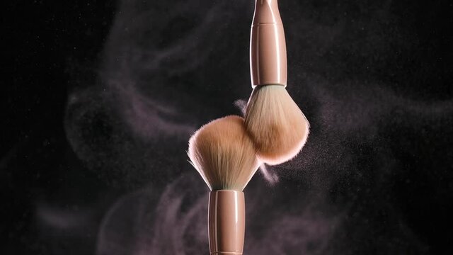 Two soft cosmetic brushes release a cloud of powder particles in contact with each other. Close up of makeup brushes on a black background. Beauty industry, decorative cosmetics concept. Slow motion.