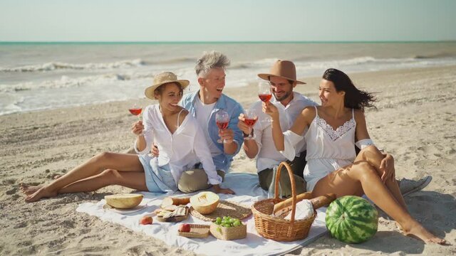 group of four happy friends celebrating with wine and having toast at breakfast at summer beach party or picnic. cheerful people clinking wine glasses and drinking, having fun at sea vacation travel