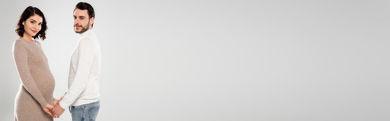 Pregnant woman holding hands of husband and smiling at camera isolated on grey, banner