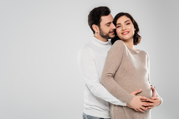 Fototapeta premium Happy man hugging cheerful pregnant woman isolated on grey