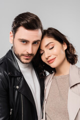 Man in leather jacket looking at camera near brunette wife isolated on grey