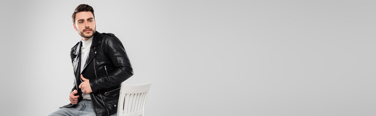 Trendy man in leather jacket sitting on chair isolated on grey, banner