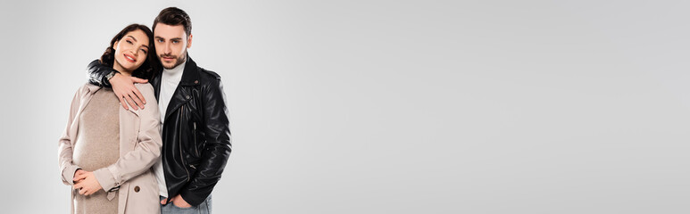 Smiling man hugging cheerful pregnant wife isolated on grey, banner