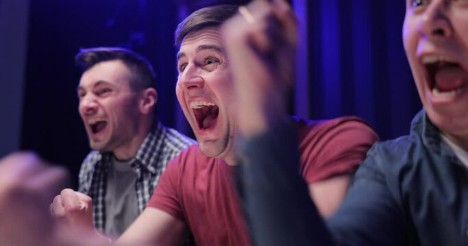 Close up of Caucasian male football fan sitting with friends at TV screen in dark living room and watching match. Cheerful man celebrating football win. Scoring goal or winning game concept