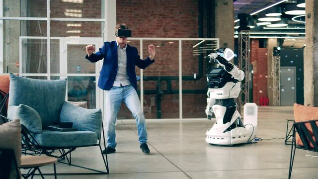 A man in VR-glasses is dancing with a modern robot