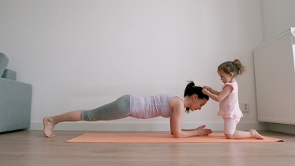 Mother in plank exercise and little girl plays with her mother's hair at home