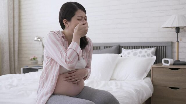 pretty Korean lady seated on bedside covering her mouth is having morning sick and wanting to puke during pregnancy at bright home interior.