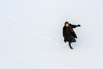 a cheerful girl in a warm fur coat walks across the open field leaving footpath in the snow