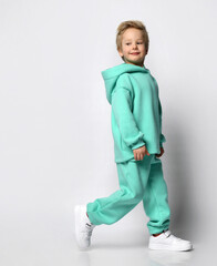 Full portrait of stylish little boy in warm tracksuit against gray background. A boy in a turquoise fleece suit and sneakers stands sideways to the camera near free space for text.