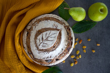 Beautifully scored rye sourdough bread with leaf pattern on the top. Round bread, raisins, green leaves and fruits on yellow fabric background.