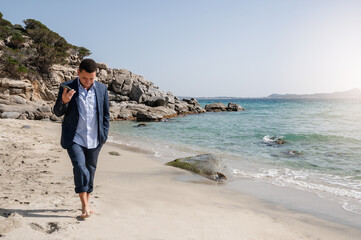 Latin businessman with suit walking on beautiful sandy beach while holding a mobile phone and...