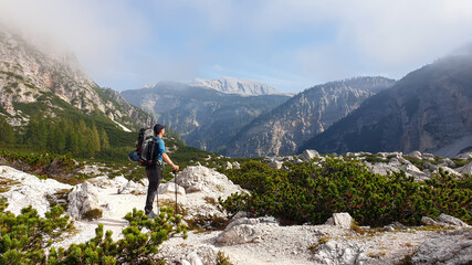 A man with a big hiking backpack enjoying the panoramic view on Italian Dolomites. He stands on a stony plateau, above the valley. A few clouds around. In the back high mountain chains. Freedom