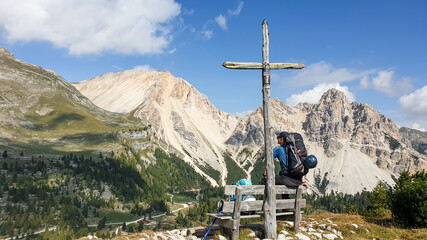 A man with a big hiking backpack sitting on a wooden bench under a wooden cross end enjoying the panoramic view on a green valley in Italian Dolomites. High and sharp mountains everywhere. Freedom