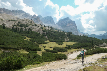 Fototapeta na wymiar A woman with a hiking backpack hiking on a gravelled road in high Italian Dolomites. There are a few trees on the lower parts, and steep and sharp mountain chain in the back. Discovering and exploring
