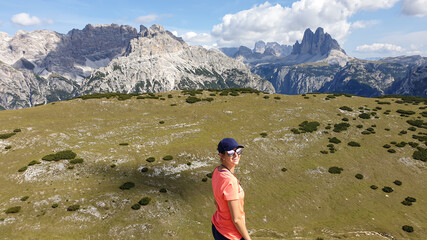 A woman in sunglasses with a distant view on Drei Zinnen from Strudelkopf in Italian Dolomites. A lush green meadow in front. The woman is smiling, enjoying her hike. High Alpine landscape. Freedom