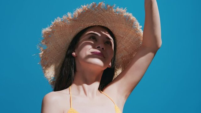 Close-up beauty portrait of young dark-haired asian woman in the straw hat covers her face from the sun with her palm turns to the camera and smiles against blue background | Sunblock commercial