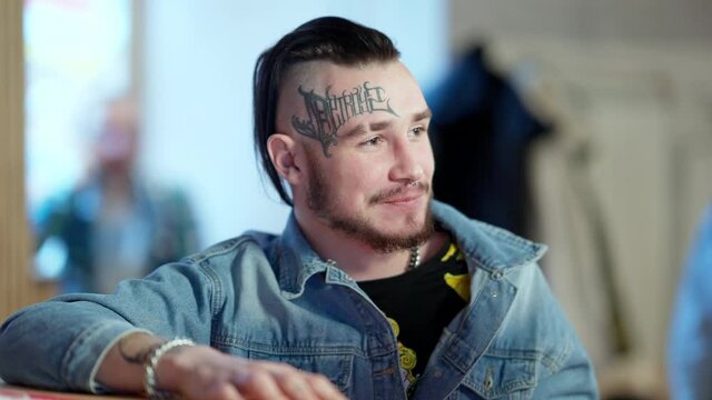 Laughing pierced young man with tattoo on face sitting at bar counter indoors. Portrait of cheerful positive Caucasian millennial resting in pub. Lifestyle and relaxation.