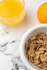 Aerial view of a glass of juice, a bowl of cereals and an orange on white marble, in vertical, with copy space
