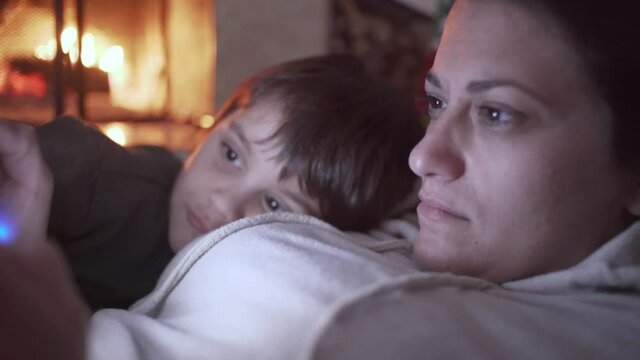 Close up shot of caucasian mother playing console video game,while here son watching and laughing besides her. Fireplace burning in the blurry background