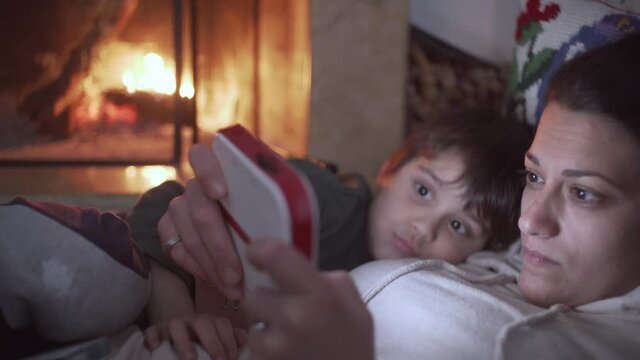 Caucasian mother and her son, relaxing by the fireplace, playing videogames on a console, pan medium shot