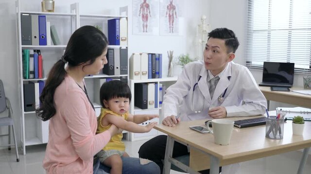 asian pediatrician is observing the young child and asking the mother questions during the appointment in his clinic office.