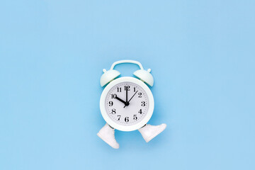 White alarm clock in toy sneakers walks on blue background. Minimal time movement concept. Place...