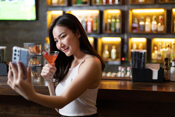 Young Asian woman takes a photo of herself or Selfie with a smiling face and drinks a cocktail in a vintage bar, Relaxing activities after work or hangouts, Place of entertainment for teens.
