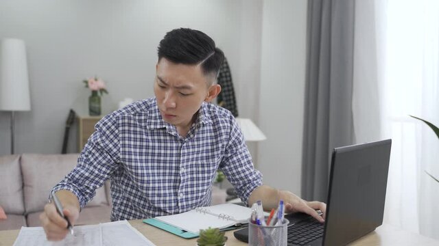 asian accountant working from home on the computer is taking notes while going through company expenses with concentration.