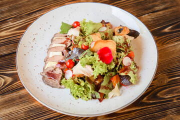 Salad with sliced meat, cheese, vegetables and mix lettuce on a white plate on a wooden table. Appetizing restaurant dish.