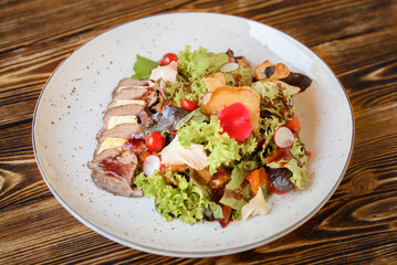 Salad with sliced meat, cheese, vegetables and mix lettuce on a white plate on a wooden table. Appetizing restaurant dish.
