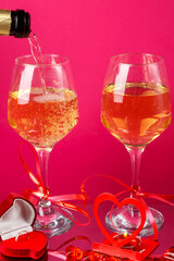 Champagne is poured into glasses on a pink background next to a heart candlestick with a burning candle and a box with a ring.