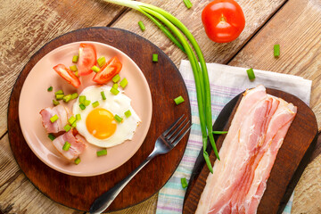 Scrambled eggs with bacon with tomato and green onions and on a wooden table in a plate on a stand with a fork next to chopped bacon and tomatoes.