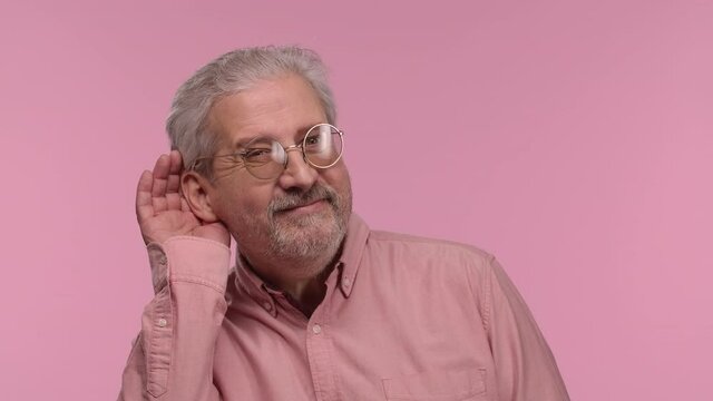 Portrait Of An Elderly Man With Glasses Holding Hand Near Ear Trying To Listen To Interesting News Expressing And Gossip. Gray Haired Pensioner Posing On Pink Studio Background. Close Up. Slow Motion.