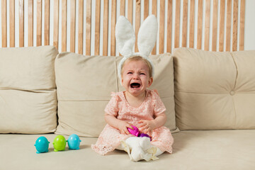 crying little girl in a headband with bunny ears with egg toys sits on a beige sofa with a basket...