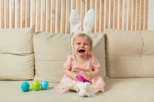 crying little girl in a headband with bunny ears with egg toys sits on a beige sofa with a basket with a cotton flower