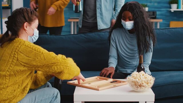 Cheerful woman using dice playing backgammon with multiethnic friends wearing face mask as prevention against covid 19 virus during global pandemic sitting on couch drinking beer and eating popcorn