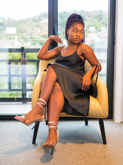 Fashion lifestyle of a black ethnic girl in a black dress. Portrait of the pretty girl sitting on a yellow chair
