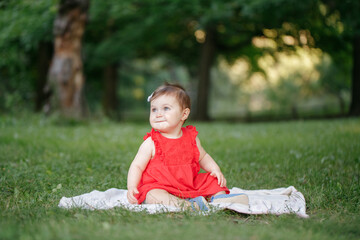 Cute adorable baby girl in red dress sitting on white blanket in park outdoor. Funny child toddler playing and having fun on summer day outside. Funny kid. Authentic lifestyle happy childhood.