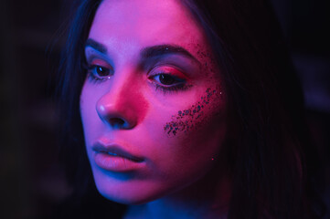 Fototapeta premium Close portrait of beautiful woman with trendy makeup in violet night light, looking away. Night photo of a brunette in neon light.